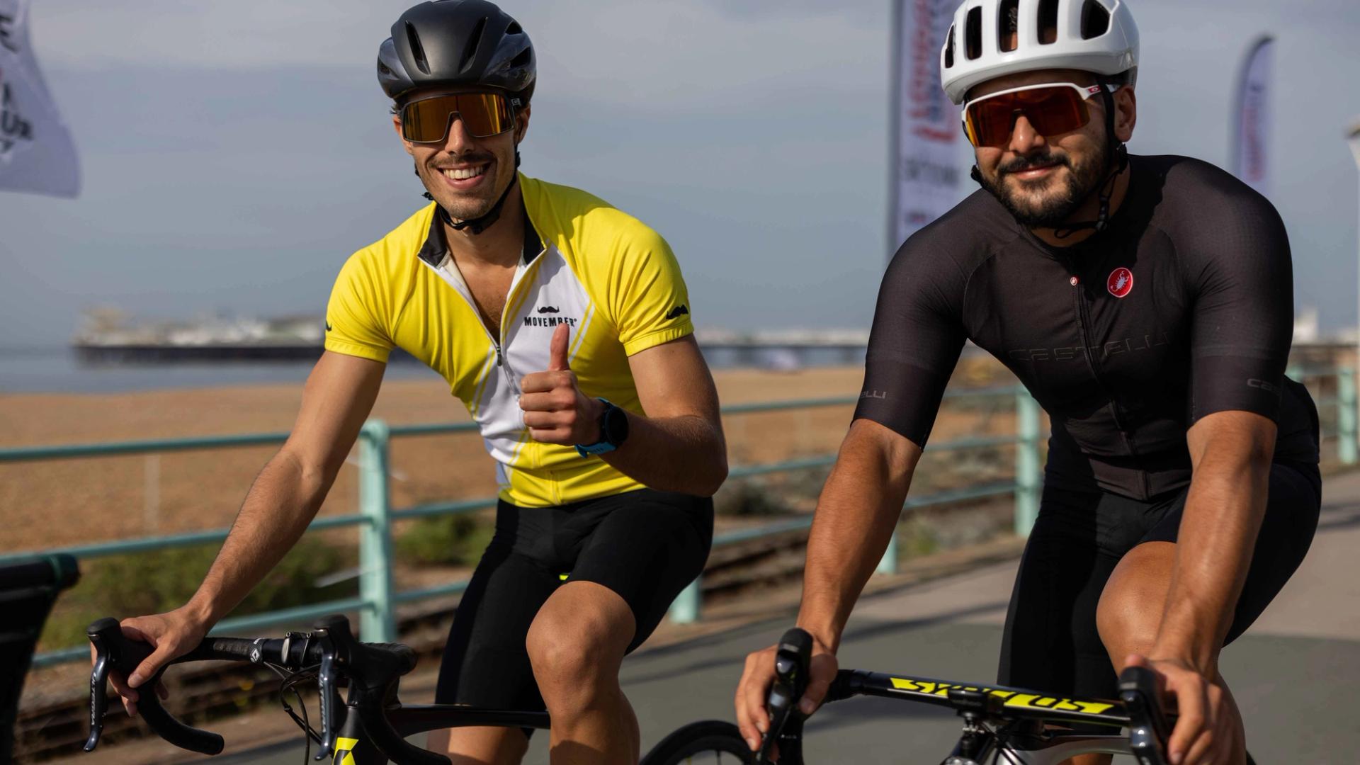 Photo of two cyclists riding bikes, clad in Movember-branded riding gear, smiling to camera.