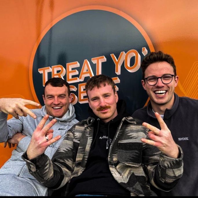 Photo of three men, smiling an looking very enthusiastically to camera. A partly obscured sign in the background, reads: "Treat Yo' Self".